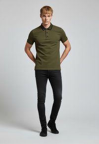 Olivově zelené polo tričko s černým vzorovaným límečkem a krátkými rukávy. Kombinováno s černými slim džíny a černými teniskami.