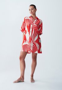 Vestito a camicia rosso e bianco con motivo, maniche corte, chiusura con bottoni e vestibilità ampia, abbinato a sandali sottili con cinturini.