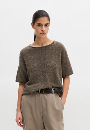 Femme aux cheveux foncés attachés porte un haut marron texturé à manches courtes, rentré dans un pantalon beige taille haute avec une ceinture noire.