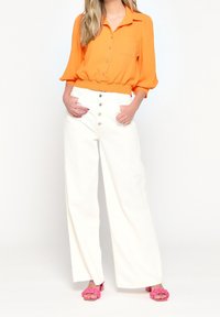 Camicia arancione con colletto e vita cintrata, abbinata a pantaloni bianchi a gamba larga con bottoni frontali e sandali rosa.