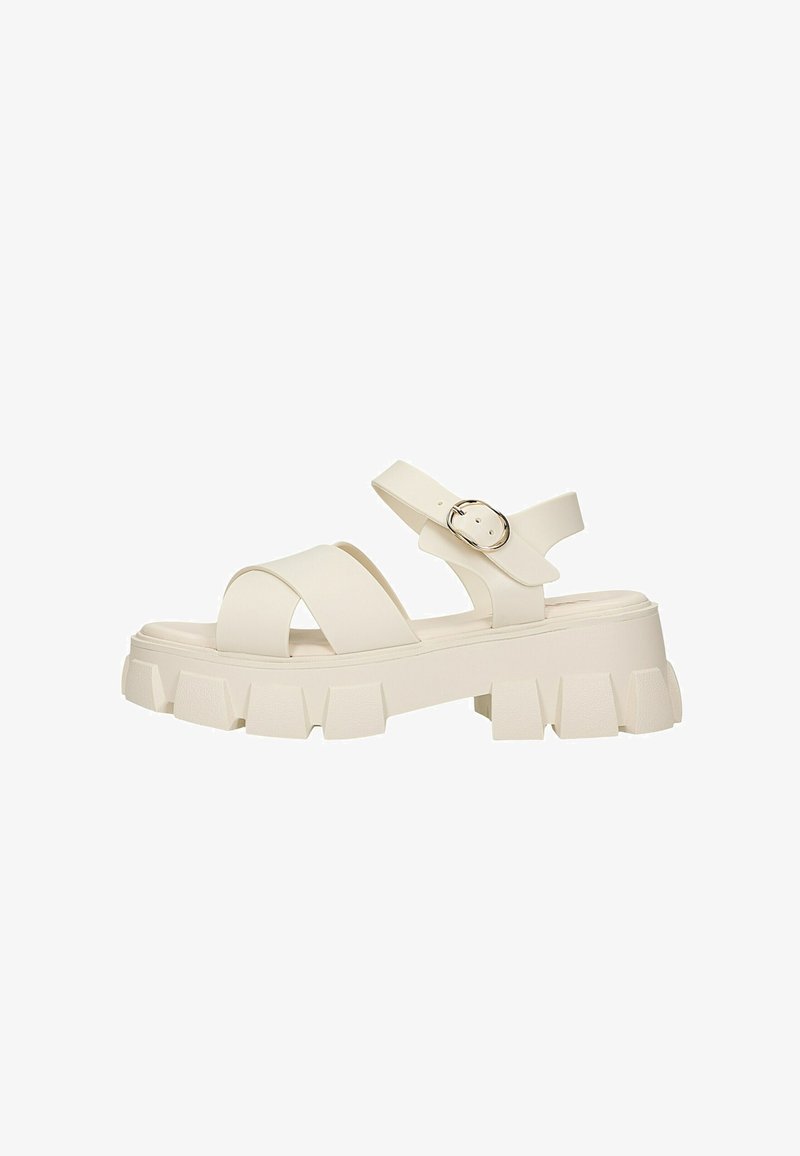 Sandale beige à semelle épaisse avec de larges brides croisées et une bride de cheville avec une boucle ronde argentée sur fond blanc.