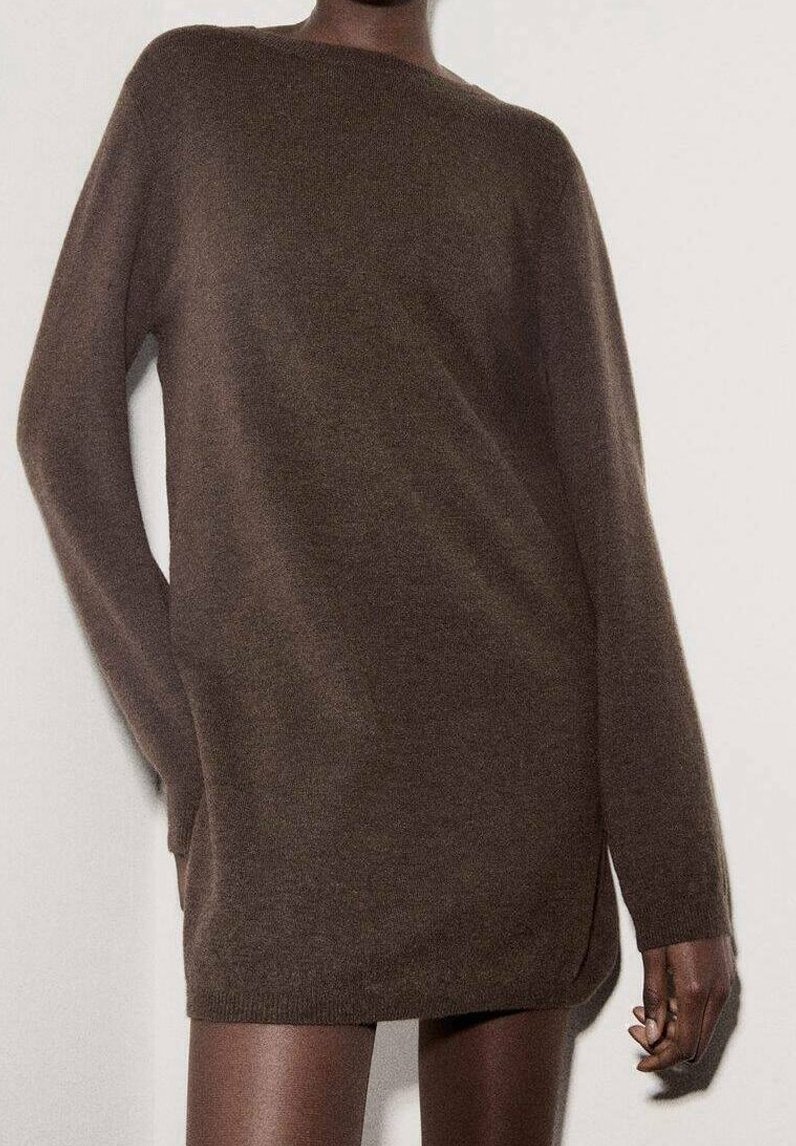 Personne portant une robe-pull marron à manches longues et oversize, se tenant contre un mur clair uni, une main détendue le long du corps.