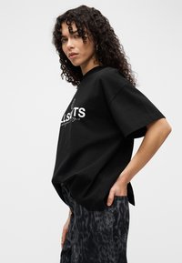 Γυναίκα με σγουρά μαλλιά, φοράει μια μαύρη υπερμεγέθη μπλούζα και σκουρόχρωμο παντελόνι με σχέδια, ποζάροντας με το ένα χέρι στην τσέπη μπροστά σε απλό φόντο.