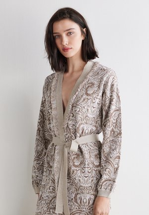 Junge Frau trägt einen beigen Bademantel mit Paisley-Muster und Gürtel, mit langen Ärmeln, stehend vor einem einfarbigen weißen Hintergrund.