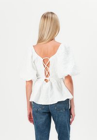 Blusa bianca strutturata con maniche a sbuffo e un design di lace-up legato sulla schiena. Abbinata a jeans blu di denim.