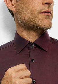 Bordeauxfarbenes Dress-Shirt mit klassischem Kragen, schwarzen Knöpfen und einer glatten Textur. Details umfassen die Nähte des Shirts und einen subtilen Glanz.