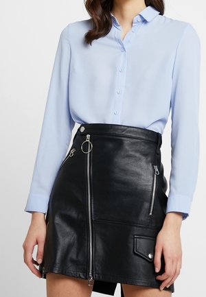 Kvinde iført en lyseblå skjorte med knapper, som er stoppet ned i en sort lædermininederdel med flere sølvlynlåse og en lomme.