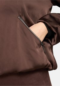 Brown pullover with a smooth, matte texture featuring a side zip pocket. The fabric is lightweight and semi-structured, with a relaxed fit.