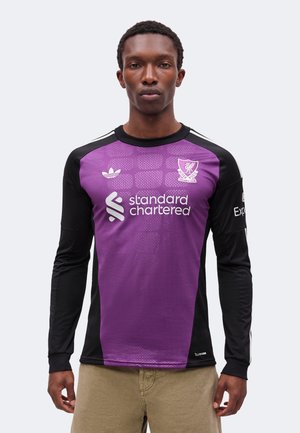 Mężczyzna noszący fioletową i czarną koszulkę z długim rękawem Liverpool Football Club z logo "standard chartered", stojący na jednolitym tle.