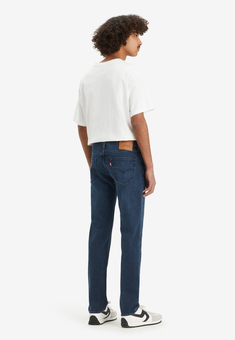 Jean bleu à coupe droite, en matériau coton, avec une patch en cuir tan à l'arrière. Porté avec un t-shirt blanc simple et des baskets.