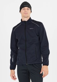 Veste softshell marine à fermeture éclair complète, col montant, accents réfléchissants et poches latérales. Portée avec un bonnet noir et un pantalon noir.