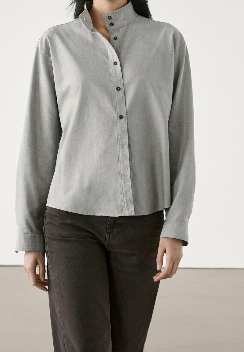 Femme portant une chemise gris clair à manches longues avec un col montant et un pantalon noir, debout devant un fond uni.