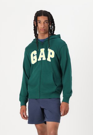 Sweat à zip vert avec un grand logo jaune "GAP" sur le devant. Il comporte une capuche à cordon et une poche kangourou. Porté avec un short marine.