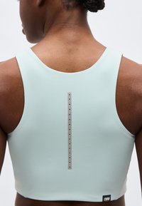 Πίσω όψη ενός ατόμου που φορά ένα ανοιχτό μπλε αθλητικό σουτιέν με κάθετη ανακλαστική λωρίδα και το λογότυπο "ciele" κοντά στο κάτω άκρο.