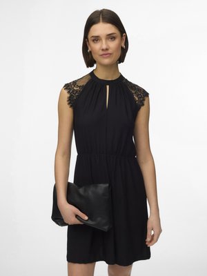 Kvinne iført en svart ermeløs kjole med blonder på skuldrene, holder en stor svart clutchveske i den ene hånden foran en ensfarget hvit bakgrunn.