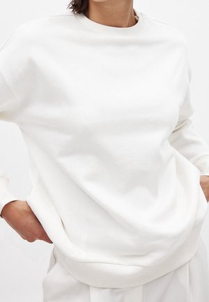 Personne portant un sweat-shirt blanc uni à manches longues, mains dans les poches, visible des épaules aux hanches sur un fond clair.
