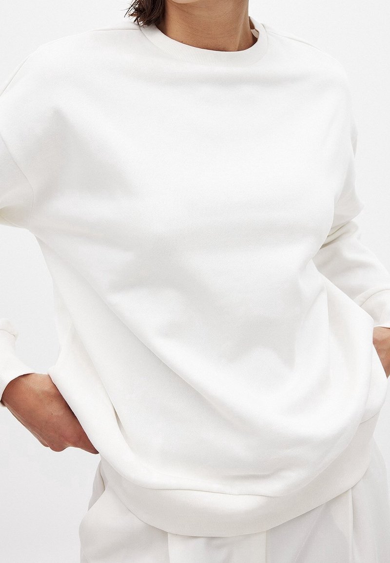 Personne portant un sweat-shirt blanc uni à manches longues, mains dans les poches, visible des épaules aux hanches sur un fond clair.