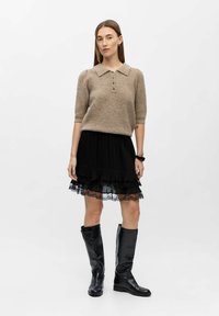 Beżowy sweter polo z dzianiny, z pięcioma guzikami, zestawiony z czarną plisowaną spódnicą z koronkowym wykończeniem oraz wysokimi czarnymi skórzanymi botkami.