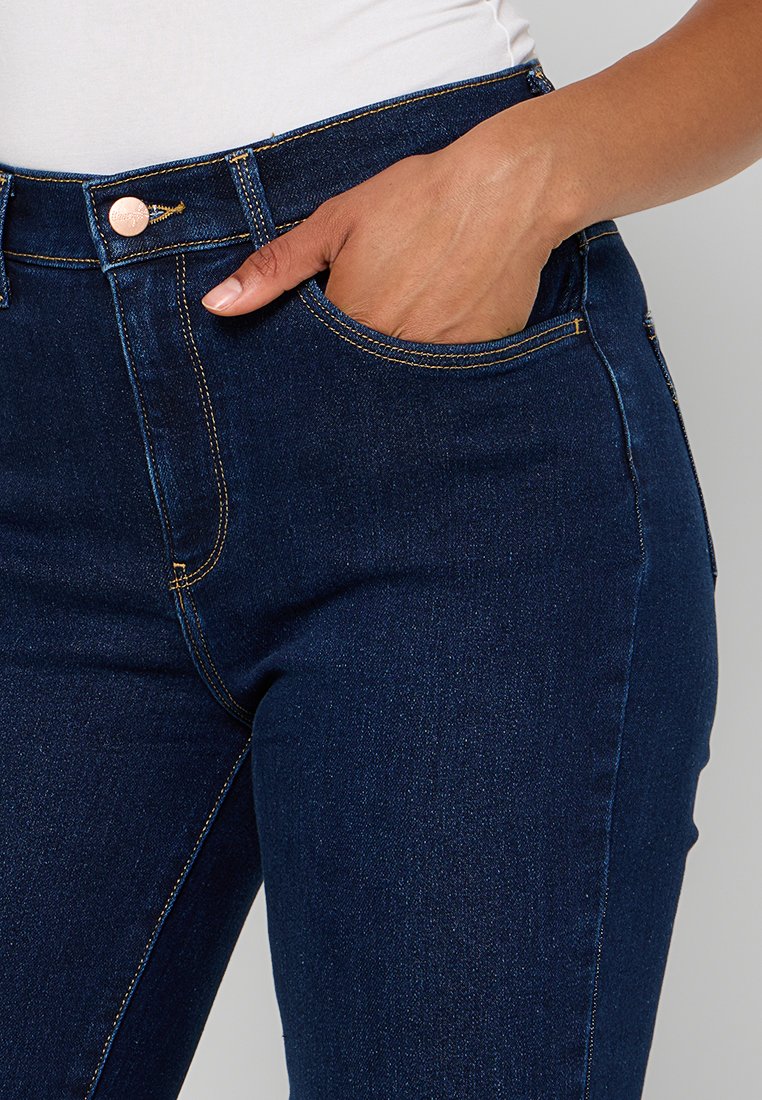 Jean en denim bleu foncé au design ajusté, doté d'une poche avant, de coutures visibles et d'un bouton en or rose à la taille.