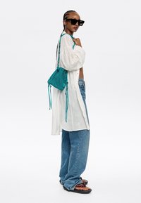 Borsa intrecciata turchese a forma triangolare, con tracolla e frange; abbinata a una camicia bianca ricamata e jeans larghi.