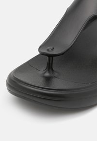 Sandale tongs en caoutchouc noir avec semelle texturée et large bride fixée par un entre-doigt noir sur un fond uni.
