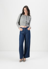 Šedý žebrovaný zkrácený cardigan s knoflíky, spárovaný s širokými tmavomodrými džínami. Modelka má na sobě špičaté šedé baleríny a nese černou tašku.