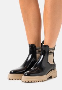 Botines de charol negro con panel lateral elástico beige, tacón texturizado y suela gruesa antideslizante, con detalle de marca en contraste.