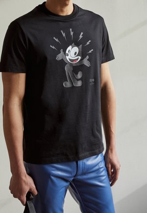 Mann trägt ein schwarzes T-Shirt mit Felix the Cat-Motiv und blaue Lederhose, steht vor einem einfarbigen hellen Hintergrund.