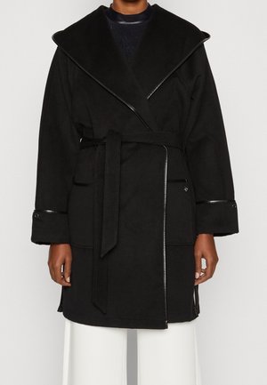 Personne portant un manteau noir ceinturé avec un large col et des détails en cuir, associé à un pantalon blanc ample.