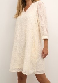 Løs, cremefarvet kjole med V-hals, trekvart gennemsigtige ærmer og blomsterbroderi over det hele. Blødt, tekstureret stof.