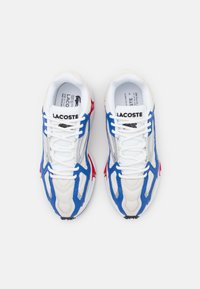 Lacoste sportcipő, amely fehér, kék és piros színben, hálós és szintetikus felsőrésszel rendelkezik, fűzős kialakítással és texturált talppal a tapadás érdekében.