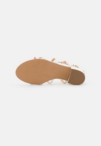 Vit sandal med flera remmar, en slät yta och en brun gummisula med lätt greppmönster och en platt design.