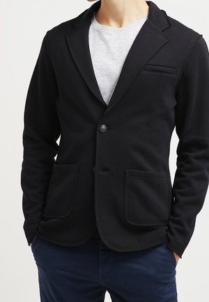 Hombre que lleva una blazer negra de un solo botón sobre una camiseta gris claro con pantalones azul marino, manos parcialmente en los bolsillos.