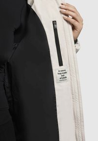 Veste blanche avec des passepoils noirs, dotée d'une poche zippée et d'une étiquette indiquant : "Un câlin chaud en tissu." Texture douce et matelassée.