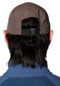 Brun keps med en texturerad tyg, justerbar svart rem bak, bärs med en blå jacka med hög krage, hår syns under.