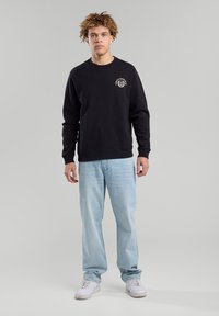 Black sweatshirt with a circular logo on the chest, paired with light blue jeans and white sneakers. Simple, casual design.