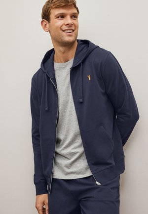 Marineblauwe ritskap met trekkoorden, voorzakken en een klein geborduurd logo. Draagt over een lichtgrijze T-shirt met een zachte textuur.