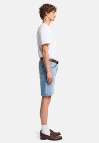Lysblå denimshorts med en rå kant, hvid bomulds t-shirt og brune læder loafers parret med hvide ankelstrømper. Sideprofilbillede.