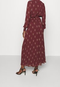 Femme portant une robe longue bordeaux à manches longues avec un petit motif floral doré, associée à des sandales noires à talons hauts, vue de dos.