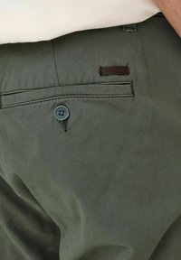 Primer plano de un bolsillo de pantalones color verde oliva con un botón y una pequeña etiqueta de cuero con la marca encima de la costura del bolsillo.