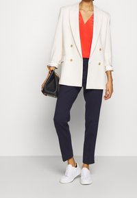 Donna che indossa un blazer bianco, una blusa rossa, pantaloni blu navy e sneaker bianche, mentre tiene una borsa nera con manici in legno, su uno sfondo neutro.
