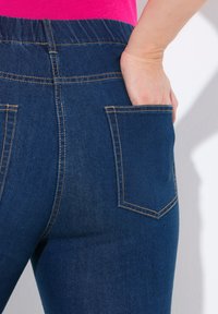 Džínové kalhoty s tmavě modrou barvou, se šitými zadními kapsami a elastickým pasem pro pohodlí. Materiál má hladkou texturu.