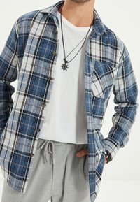 Camicia di flanella a quadri in blu e bianco, con chiusura frontale con bottoni, taschino sul petto, maniche lunghe e vestibilità comoda. Indossata sopra una t-shirt bianca.
