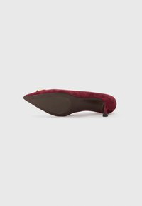 Scarpa con tacco alto in suede color bordeaux, con punta affusolata e finitura in tessuto testurizzato. Presenta un tacco sottile e una suola liscia marrone.