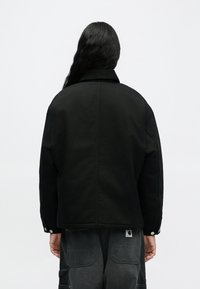 Osoba s dlhými tmavými vlasmi, otočená chrbtom, oblečená v čiernej bunde a tmavosivých nohaviciach s nášivkovými vreckami, na jednoduchom pozadí.