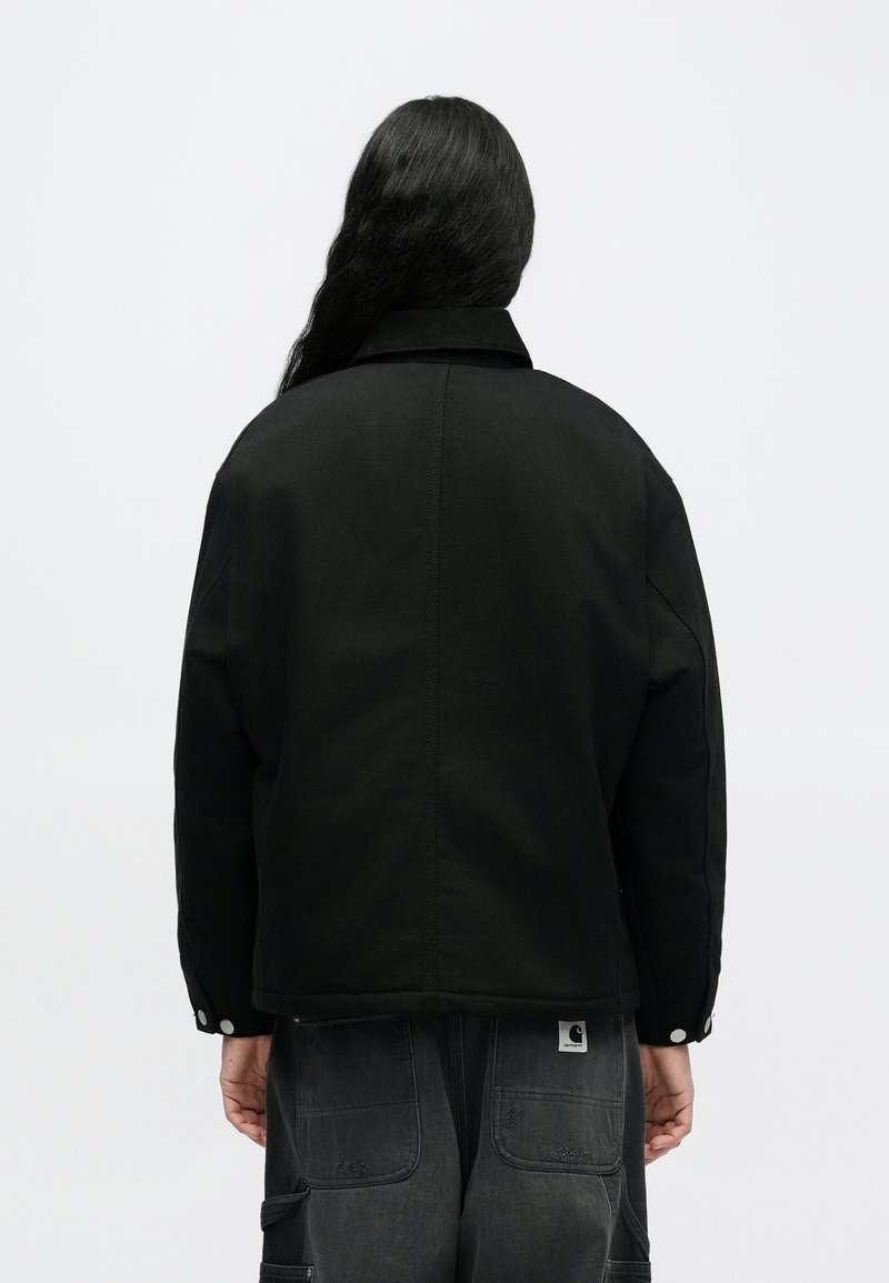 Persona con cabello largo y oscuro vista de espaldas, vistiendo una chaqueta negra y pantalones gris oscuro con bolsillos parche contra un fondo liso.