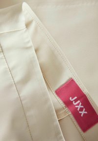 Beige stofdetail met een gladde textuur, vergezeld van een roze label met witte tekst "JJXX." Stiksel is zichtbaar langs de rand.