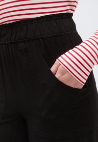 Μαύρο παντελόνι με ελαστική μέση και πλαϊνές τσέπες. Η υφή είναι ελαφρώς ζαρωμένη. Μερικώς ορατό ένα μακρυμάνικο πουκάμισο με λευκές και κόκκινες ρίγες.