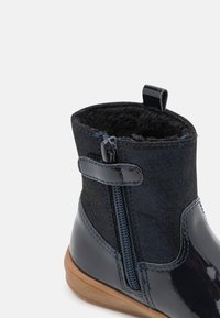 Botte cheville bleu marine avec une finition brillante en vernis, doublure intérieure douce, fermeture éclair sur le côté et un panneau en tissu texturé. Semelle en caoutchouc avec une légère adhérence.