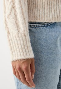 Main gauche reposant à côté d'un jean en denim bleu clair, portant un pull en maille côtelée couleur crème avec des motifs torsadés sur la manche.
